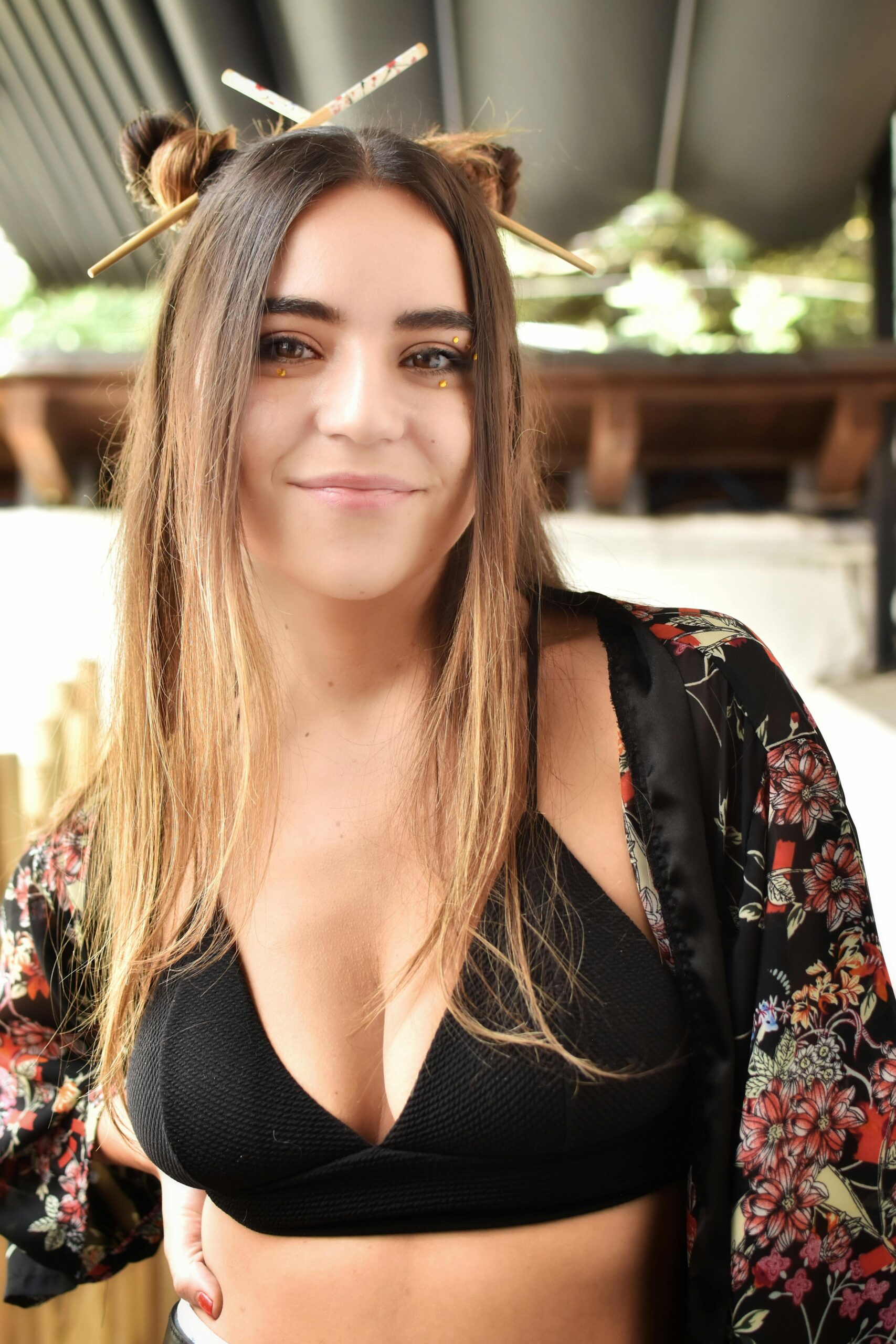 Smiling young woman with chopstick hair buns in fashionable black top and floral kimono outdoors.
